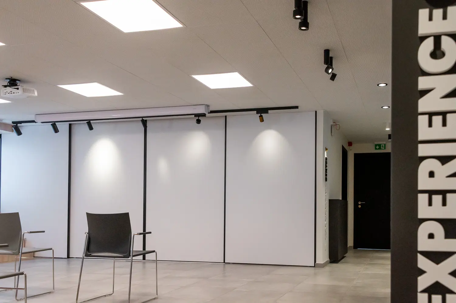 Vue intérieure d'un espace de bureau moderne avec des dalles d'éclairage carrées, des chaises de bureau empilées et de grands panneaux blancs, avec le mot "EXPERIENCE" vertical sur un mur